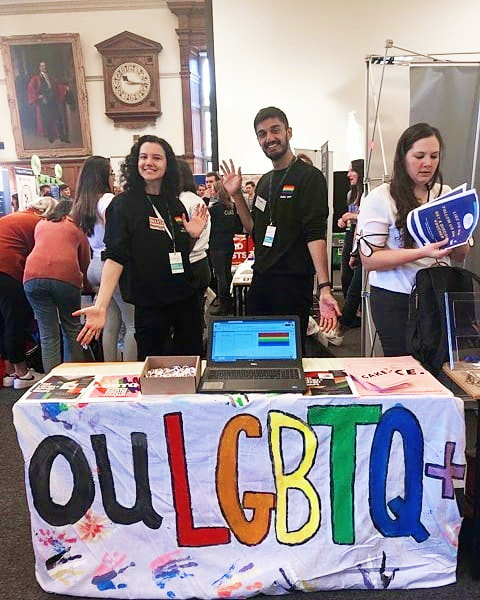 A photo of the OULGBTQ+ Society stand at the Freshers' fair. A sheet with OULGBTQ+ written on it in big rainbow writing is placed over the stall with 2 committee members smiling into camera behind.