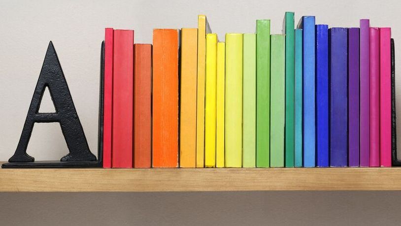 A photo of a book case, with each book a different colour making a rainbow. The letter 'A' props them up.