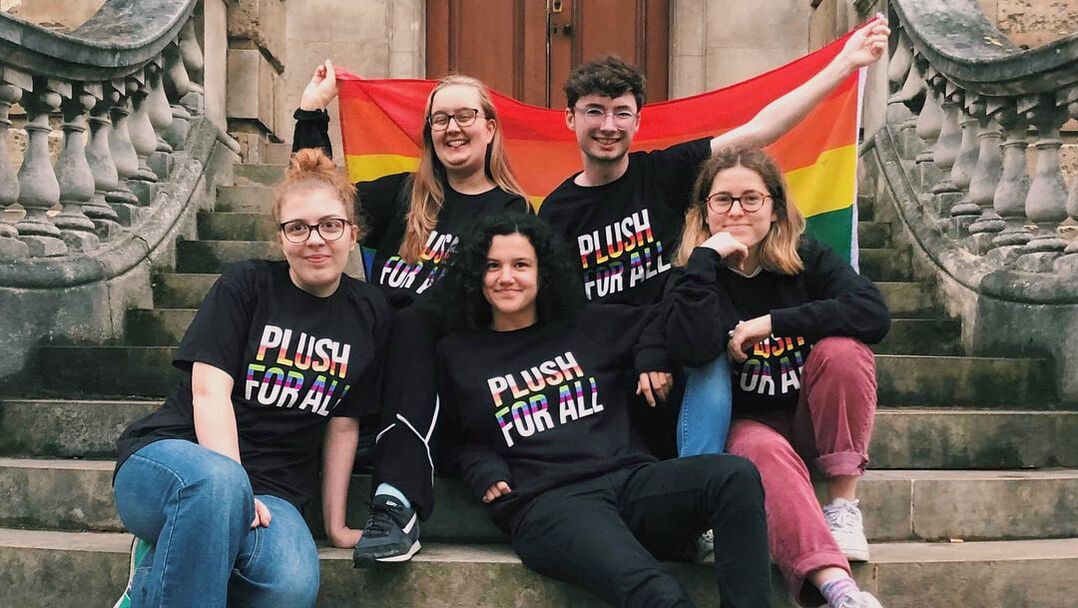 A few of the oulgbtq+ society committee sat on the Radcliffe Camera steps holding a Pride Flag behind them and wearing black t-shirts which read 'Plush For All' in rainbow writing.