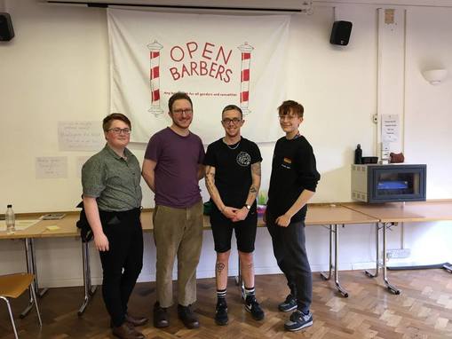 Two members of LGBTQ+ Society standing with the hairdressers Felix and Graham in front of the red and white Open Barbers flag.
