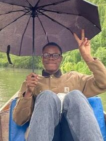 Mbere, who has short dark hair and is wearing glasses, makes a peace sign. He is sat on a boat holding an umbrella